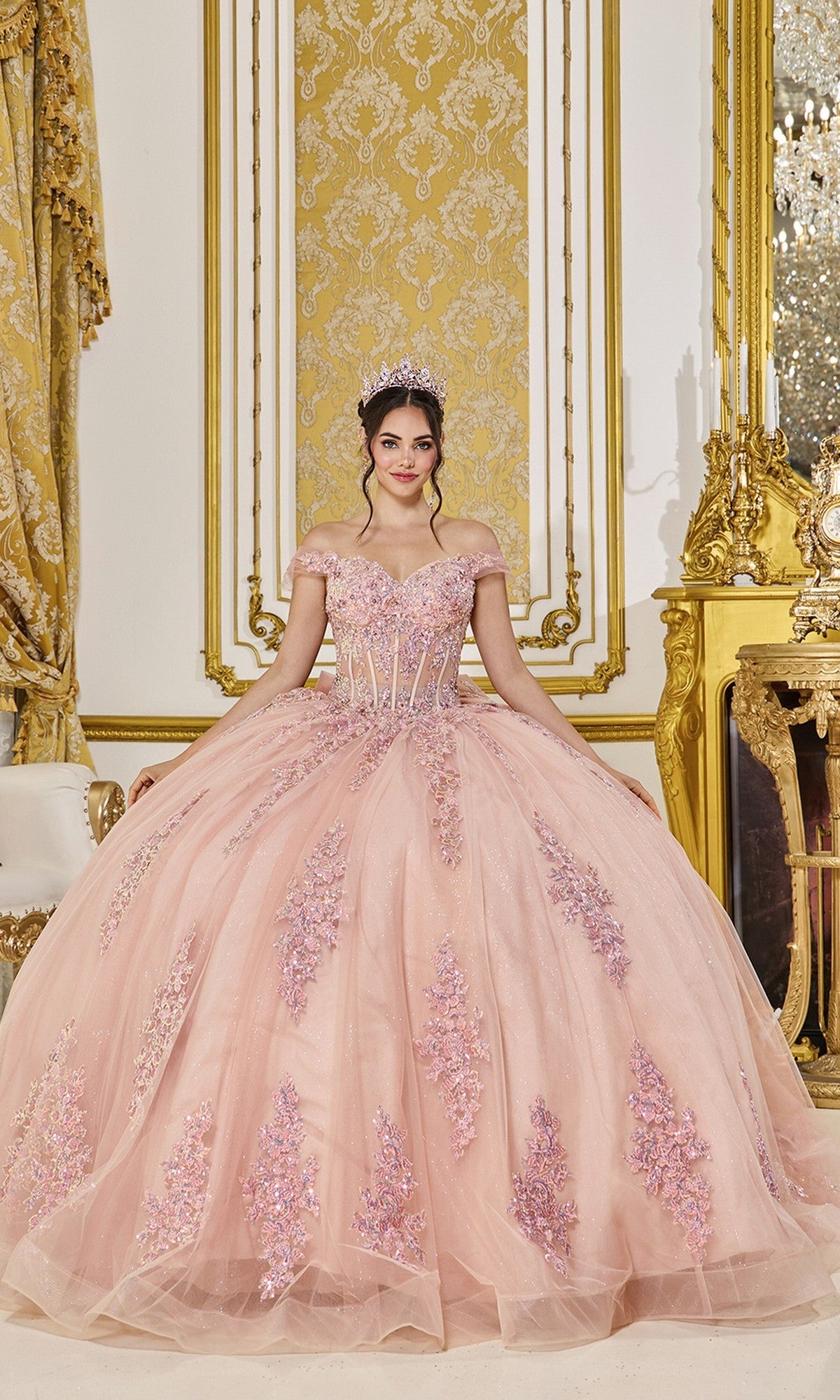 A young woman wears the Quinceanera Dress 5121 By Dancing Queen in rose gold, featuring floral lace appliqué and an off-the-shoulder neckline, as she stands in a lavish gold-decorated room with a sparkling tiara above her embellished ball gown.